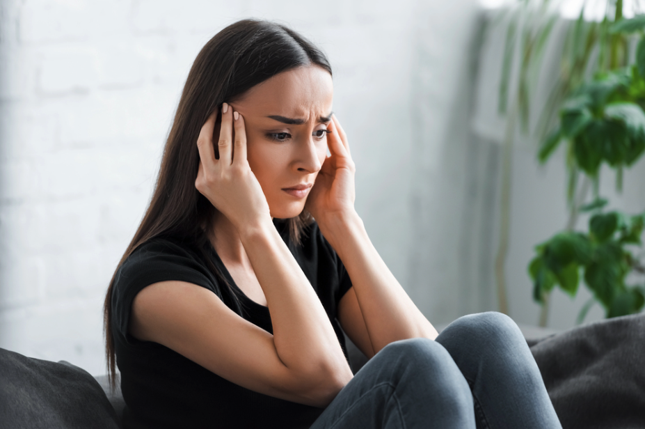 A woman sitting on a sofa, holding her head with a worried expression, indicating stress or headache.