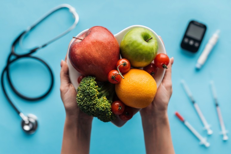 Hands holding a bowl of fresh fruits and vegetables (apple, orange, broccoli, tomatoes) with medical tools in the background.
