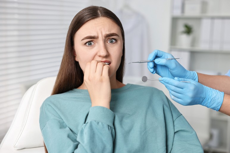 Woman in a dental chair looks anxious as a dentist's gloved hands hold dental tools near her mouth.