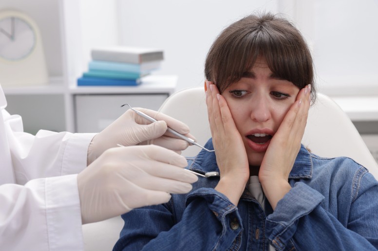 An anxious woman sits in a dental chair with a clinician’s gloved hands holding dental tools beside her open mouth.