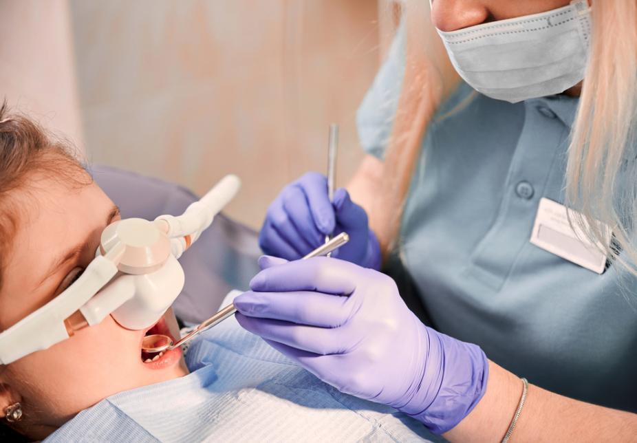 march26c Dentist wearing a mask and purple gloves works on a patient's teeth with dental instruments in a reclining chair.