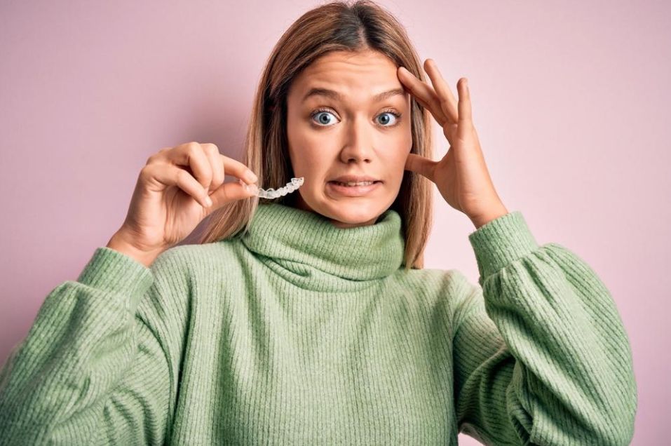 june-a Woman in a green sweater holding a small dental appliance fragment, looking anxious against a pink background.