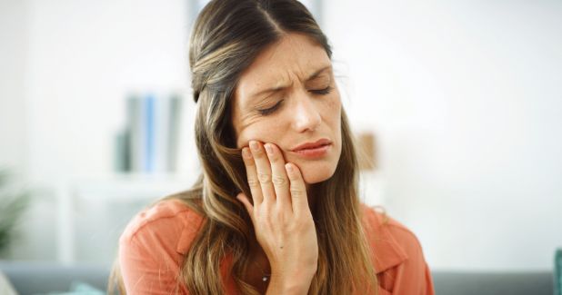 may26c Woman grimacing in pain while holding her cheek, suggesting a toothache or jaw pain.